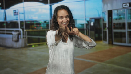 Fototapeta premium Woman framing with hands and pointing gesture at airport entrance in white blouse, smiling and engaging camera; playful confidence.