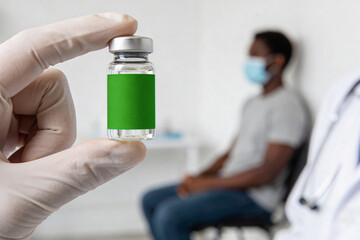 Obraz premium Doctor holding blank vaccine ampoule in front of African American patient wearing medical mask. Development of remedy against Ebola and measles. Healthcare and immunization concept.