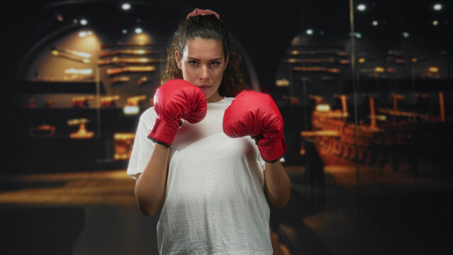 Woman in white t shirt with red boxing gloves holds fists up, standing center with intense gaze facing camera in studio; determination.