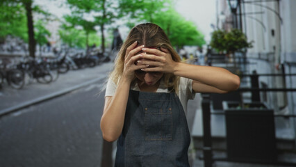 Fototapeta premium Woman with hands on head holding forehead and closed eyes on a cobblestone street near parked bicycles and planters; stress coping.