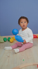 Naklejka premium Little girl wearing white shirt and pink leggings puts a blue ball to her mouth while sitting on a wooden floor surrounded by colorful toy pins in a studio; curiosity discovery play.
