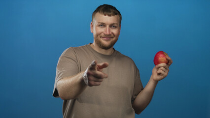 Naklejka premium Man holding red apple while pointing finger and smiling in blue studio; friendly healthy snack promotion.