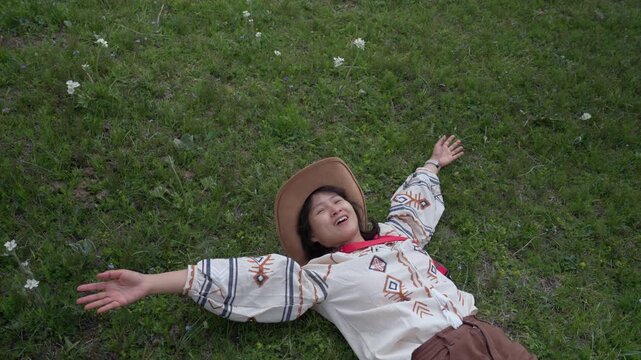 Woman with hat smile on grass meadow outdoor relax travel, joyful adult lying with arms outstretched on green field, enjoying nature under open sky, carefree freedom rest, rustic embroidery shirt vibe