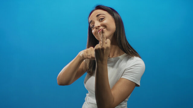 Woman shows middle finger to camera in blue studio, smiling and leaning forward in white tshirt; playful defiance.