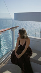 Obraz premium Woman sitting with clasped hands on cruise terrace bench overlooking ocean wake and glass railing, wearing dark dress and necklace; serenity.