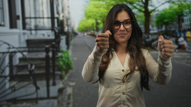 Young hispanic woman wearing glasses miming driving with hands gripping an invisible steering wheel while smiling on a city street; confidence.