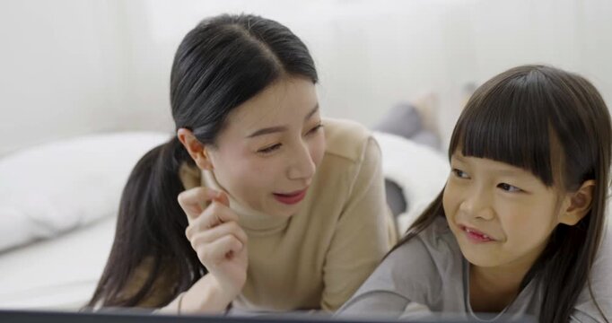 Young happy woman with cute little daughter looking at laptop computer playing on the bed, enjoying technology network together at home bedroom, happy family mothers day, education technology concept