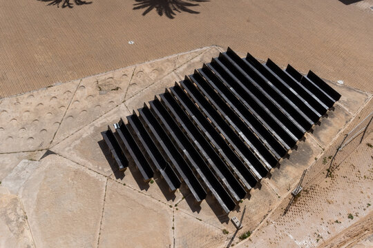 High angle view of rows of dark solar panels or parallel structures on a concrete installation in a sunny, arid or coastal landscape.