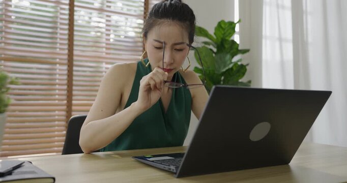 Asian business girl wearing glasses. Upset woman sitting at desk with laptop, rubbing her eyes in frustration, representing work stress, burnout, fatigue, and emotional pressure.