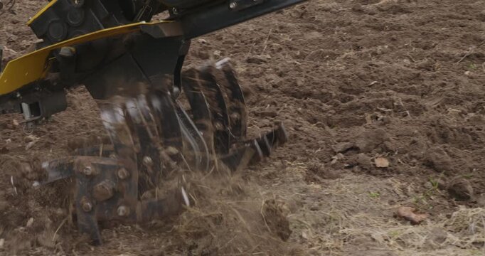 Closeup of a cultivator machine tilling the soil, preparing the garden bed for planting seeds and vegetables in the springtime, with spinning blades churning and loosening the fertile earth.