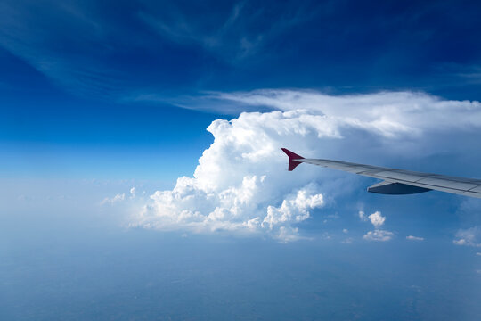 An airplane wing with a red tip extends over a vast sea of white cumulus clouds, bathed in bright sunlight under a clear blue sky during a daytime flight