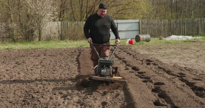 Young farmer plowing the land in his garden with a rotary tiller, cultivating the fertile soil and preparing the field for the spring planting season using modern agricultural machinery