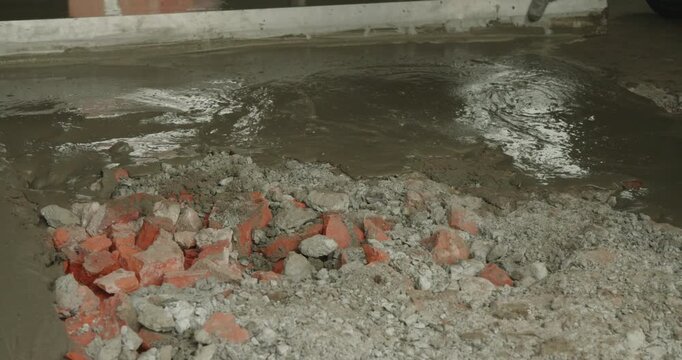 Close-up of a builder's hands pouring fresh liquid concrete from a bucket onto rubble to create a new floor, showcasing the initial stages of foundation work at a construction site