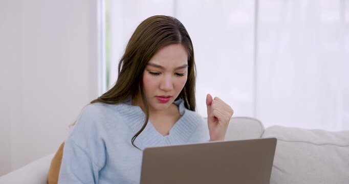 Asian young woman close up works on laptop, rests chin on hand, types, gestures slightly, reads screen with calm expression in bright apartment during relaxed home office productivity routine