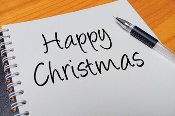 Obraz premium Top view of an open spiral notebook with Happy Christmas handwritten text overlay next to a black gel pen on a wooden desk