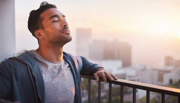 Young man enjoying a peaceful moment while standing on a balcony, breathing deeply and soaking in the sunrise over a city skyline at dawn