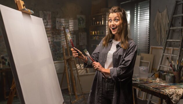 Excited Female Artist in Bright Studio Holding Paintbrushes Ready for Creative Painting on Blank Canvas with Colorful Art Supplies Around