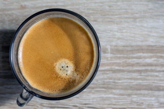 Top view of a glass cup filled with hot espresso coffee showing rich crema on a wooden table