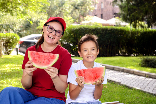 Smiling mother and son holding fresh watermelon slices in a sunny park. Healthy family lifestyle, summer refreshment, outdoor recreation, and joyful parent-child bonding concept