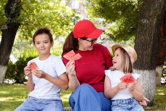 Mother and two children enjoying watermelon together in a sunny park. Happy family bonding, healthy summer snack, outdoor recreation, childhood joy, and wholesome lifestyle concept