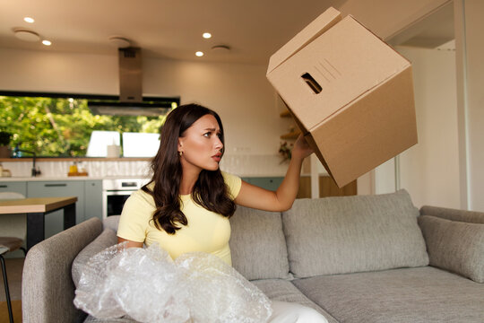 Woman sitting on sofa and looking inside empty box after opening delivery package at home, looks disappointed while holding bubble wrap