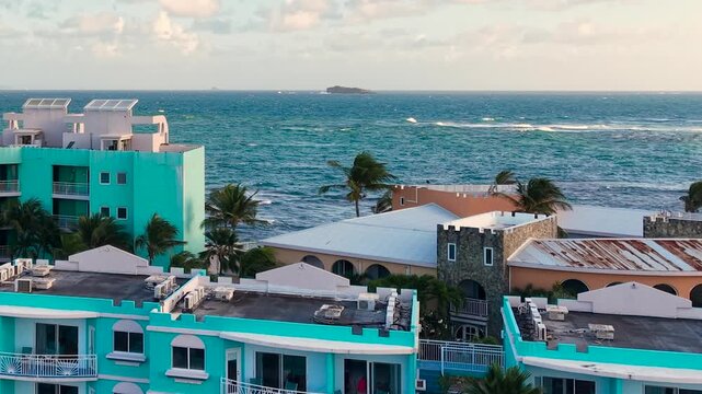 Aerial view of a coastal tropical resort on the eastern coast of Saint Martin island in the Caribbean Sea. Modern hotel buildings feature distinct white and blue architectural designs near the water.