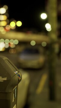 A parking meter is next to the street at night. Cars are parked in the background, and streetlights create a soft glow around the area. The scene shows city life after dark.
