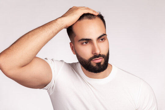 Confident man wearing white t-shirt running hand through short neat hair and showing facial contours with groomed beard masculine grooming hair style self care isolated over gray background
