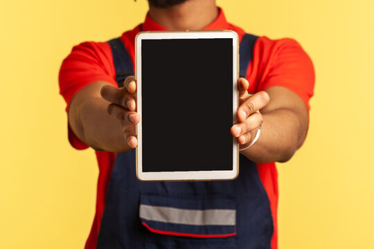 Happy handyman wearing blue overalls holding out showing tablet with blank screen with copy space for promotion, advertising industry service. Indoor studio shot isolated on yellow background.