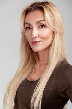 Close up portrait of beautiful blonde woman looking at camera with soft smile wearing brown long sleeve shirt standing isolated over gray background