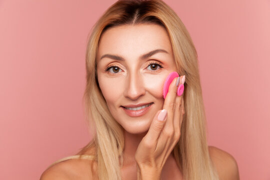 Middle aged woman using pink exfoliating pad on her cheek for professional skin cleansing and lifting isolated on pink background
