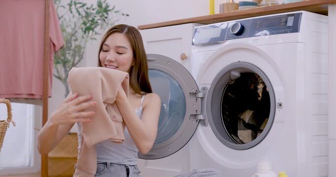 Young Asian woman checks plaid garment beside open washing machine, lifts fabric from pile, loads clothing into drum, keeping organized laundry routine in bright cozy apartment room today indoors