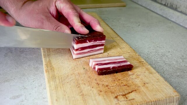 Male hands using a sharp knife to slice a piece of cured pork belly on a wooden cutting board