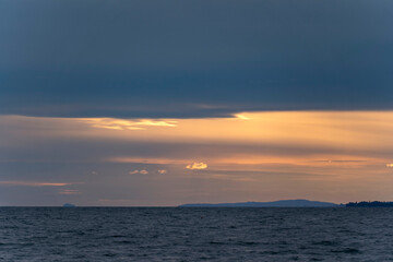 Fototapeta premium A minimalist view of Lake Constance from Bregenz at dusk, showing subtle golden sunlight breaking through dark storm clouds above wavy, deep blue waters with small illuminated clouds near the