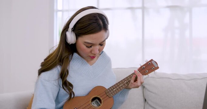 Young Asian woman plays ukulele on sofa, studies finger position, strums melody, pauses briefly, touches face, resumes music practice during relaxing weekend self care routine in bright cozy home