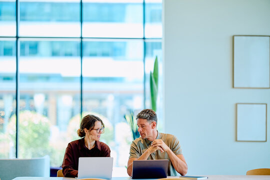 Two business colleagues discussing project ideas, working together at a table with laptops in a bright, contemporary office space