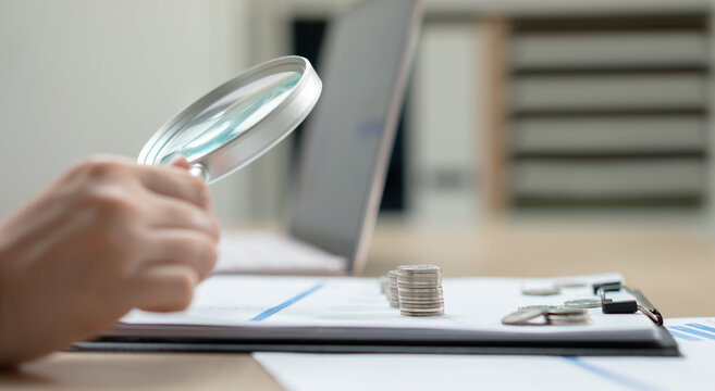 Business woman using a magnifying glass to examine financial reports and stacked coins on a desk, symbolizing audit, investment analysis, accounting, risk assessment, and business finance.