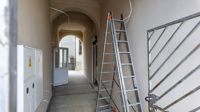 Interior of a renovated, bright tenement house corridor with arched ceilings, an aluminum stepladder leaning against the wall, white electrical boxes, and an open door leading to a courtyard.