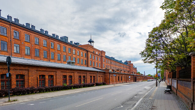 A long, historic red brick factory and former industrial complex Ksiezy Mlyn in Lodz, standing along an asphalt street with a sidewalk, trees, and a bench under a cloudy sky.