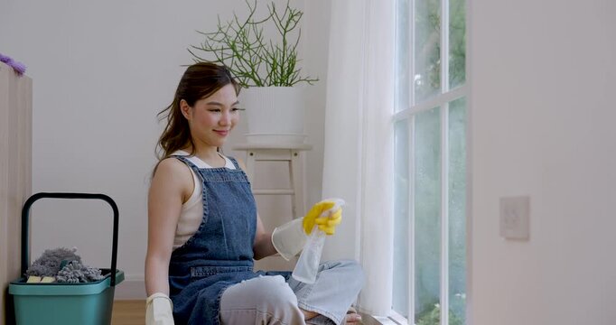 Young Asian woman sits beside window with cleaning cloth and spray bottle, leans forward to wipe glass, smiling during relaxed housework routine in bright tidy apartment interior today indoors