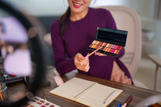 Young Asian female podcaster presenting colorful eyeshadow palette to camera while recording beauty tutorial with professional equipment in bright modern home