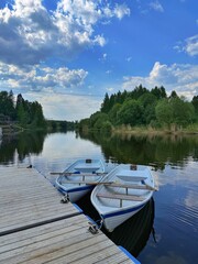 Obraz premium Two white rowboats with wooden oars are moored to a wooden pier on a calm river surrounded by forest under a cloudy blue sky. Concept of summer vacation and outdoor leisure.