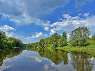 Obraz premium A calm, wide river perfectly reflects the green trees along its banks under a bright blue sky with fluffy white clouds. Concept of pristine nature and tranquil summer landscape.