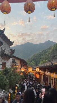 Wangxian Valley, China, 04 April 2026 ? Crowded ancient street lined with glowing red lanterns and traditional architecture frames lush green mountains at dusk in spring.