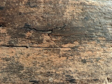 close-up view of an old, weathered wooden surface with a rich, dark brown color and prominent rustic textures. The wood features distinct horizontal cracks, deep grain lines, and irregular dark stains