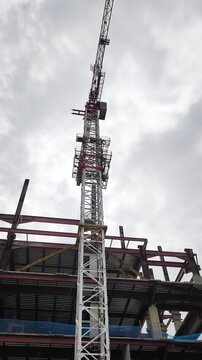 Tower crane over skeletal steel structure. A low-angle perspective captures a massive  crane pivoting against an overcast sky, operating over a building under construction. Video transition, 4k, 60 fp