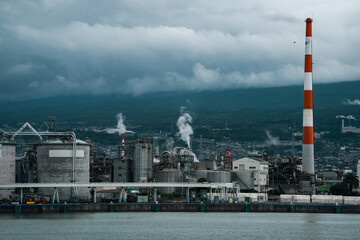 Fototapeta premium Industrial plant smokestack smoke factory refinery harbor storm cloud pollution chimney Industrial plant with smokestack and smoke near harbor under storm cloud over mountain