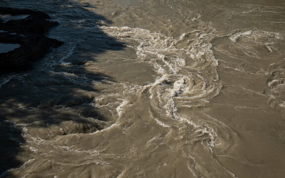 Aerial shot captures the texture of rushing, silt-heavy brown water with swirling eddy currents