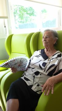 Elderly woman relaxing in a bright green armchair by a window in a nursing home. Calm senior patient fanning herself in slow motion