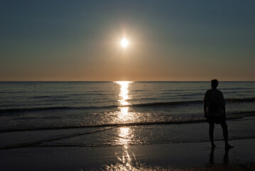 Fototapeta premium Silhouette of a person standing on a beach at sunset, watching calm ocean waves and shimmering reflections, conveying peace, solitude, travel, and mindfulness.